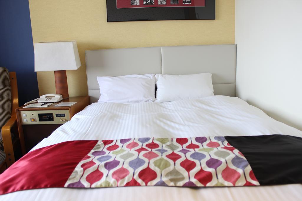 Kobe Portopia Hotel Exteriér fotografie The photo shows a neatly made bed with white bedding and two pillows. At the foot of the bed, there is a decorative blanket featuring a pattern of colorful shapes in various hues. To the side of the bed, there is a bedside table with a lamp, a teleph