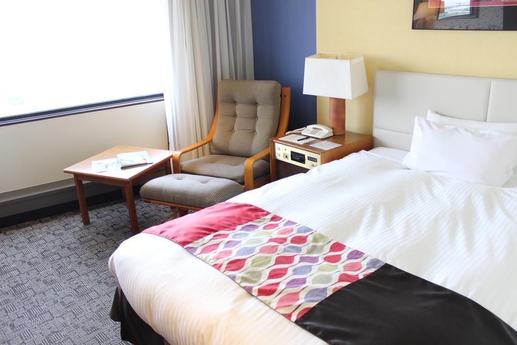 Kobe Portopia Hotel Exteriér fotografie The photo shows a comfortable hotel room. There's a bed with a white comforter and a decorative blanket featuring geometric patterns at the foot. To the side, there’s a small table with a lamp and a phone. Next to it is a cozy armchair. A window lets