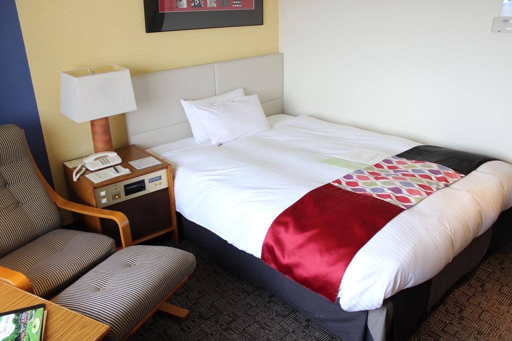 Kobe Portopia Hotel Exteriér fotografie The photo shows a small hotel room featuring a neatly made bed with a colorful throw across the bottom. Next to the bed, there is a nightstand with a lamp and a phone. In the corner, there's a comfortable armchair positioned beside a wooden side tabl