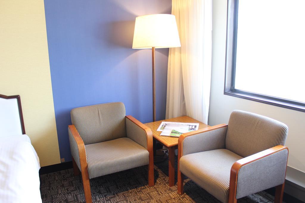 Kobe Portopia Hotel Exteriér fotografie The photo shows a small corner of a room featuring two upholstered chairs with wooden arms. These chairs are positioned around a small wooden table. Next to one of the chairs is a lamp with a white shade providing light to the area. The wall behind t