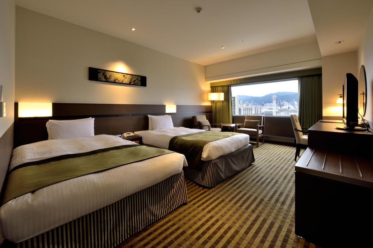 Kobe Portopia Hotel Exteriér fotografie The photo shows an interior of a hotel room. There are two beds with white linens and dark green throws. A large window offers a view of the outside, and there is a small table and chair positioned nearby. The room features warm lighting, wooden furn
