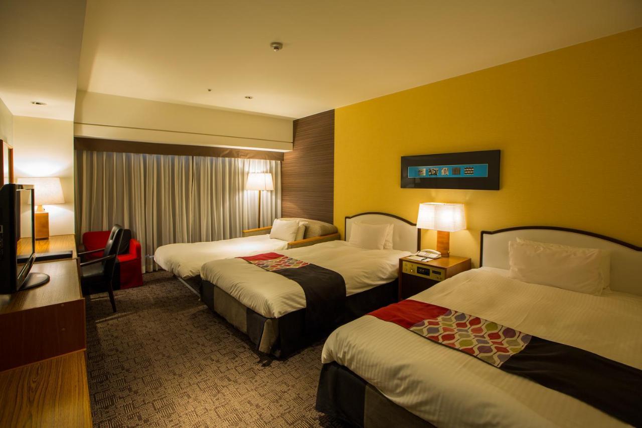 Kobe Portopia Hotel Exteriér fotografie The photo shows a hotel room featuring three beds arranged in two separate areas. Each bed has a combination of white bedding and colorful accents. There are bedside lamps providing soft lighting on either side of the beds. A large window, dressed wi
